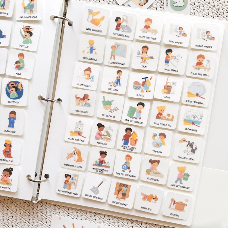 May include: A white binder filled with square cards featuring colorful illustrations of daily activities. The cards include images and text such as "Dad Comes Home," "Feed Animals," and "Take Out Garbage." The binder is open, displaying multiple rows of the cards.
