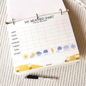 May include: A white weather chart notebook with a grid for daily observations. The title "MY WEATHER CHART" is at the top, with weather icons and a yellow floral border. A black marker pen rests below the notebook.
