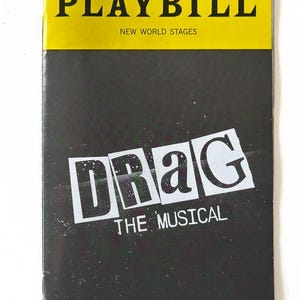 May include: A black and white playbill for the musical "Drag" with the title in bold white letters. The playbill is for New World Stages. The text at the bottom reads "City Dog Designs In partnership with Doggone Gaming."