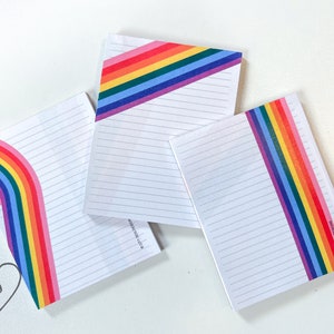 May include: Three lined notepads with rainbow designs. The notepads have a white background with black lines. The rainbow designs are on the top and bottom of the notepads. The notepads are on a white surface.