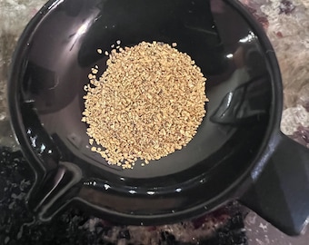 May include: A black ceramic mortar with a small pile of tan-colored grains. The mortar has a rounded bowl shape with a handle on one side. The grains appear to be spices or herbs.