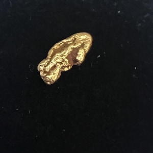 May include: A small, irregularly shaped gold nugget with a rough, textured surface. The nugget has a bright, metallic gold color and appears to be naturally formed. The background is a dark, solid color.