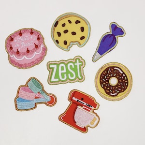 May include: Set of seven embroidered iron-on patches featuring various baking-themed designs, including a pink cake with the text "Zest is Best", a chocolate chip cookie, a purple piping bag, a green patch with the word "zest", a donut with chocolate frosting and sprinkles, a set of measuring cups, and a red stand mixer.