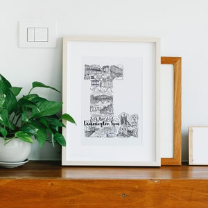 May include: A framed black and white print depicting Leamington Spa landmarks. The print is in a white frame, displayed with other framed art and a potted plant on a wooden surface. The text "Leamington Spa" is visible.