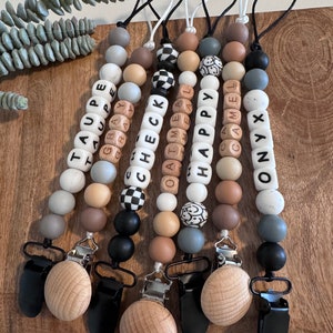 May include: A collection of baby pacifier clips with wooden and silicone beads in neutral colors. Each clip features a different word spelled out with lettered beads, including "TAUPE", "CHECK", and "HAPPY". The clips have wooden and black metal clasps.