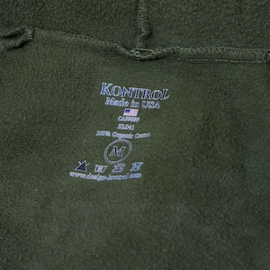 Puede incluir: Una etiqueta verde con texto blanco que dice "KONTROL Made in USA CASASES KLO41 80% Organic Cotton" y una talla "M" en un círculo. Hay un pequeño gráfico de la bandera estadounidense.