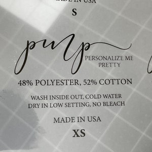 Puede incluir: Una etiqueta blanca con texto negro, que muestra las instrucciones de cuidado de una prenda. La etiqueta dice "Lavar del revés, agua fría. Secar a baja temperatura, sin blanquear. Hecho en EE. UU. XS. 48% Poliéster, 52% Algodón. Personalízame Pretty."