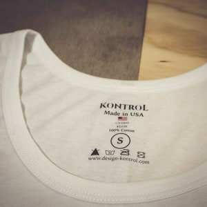 Puede incluir: Camiseta blanca de tirantes con una etiqueta negra que dice "Kontrol Made in USA" con una pequeña bandera estadounidense, "CA5995 KL018 100% Algodón", talla "S", y "www.design-kontrol.com".