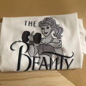 Puede incluir: Camiseta blanca con una ilustración en blanco y negro de una mujer levantando pesas. El texto "THE BEAUTY" está escrito en una fuente estilizada debajo de la ilustración.