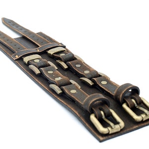 May include: A brown leather cuff bracelet with two rows of buckles and studs. The buckles are a brass color and the studs are silver.