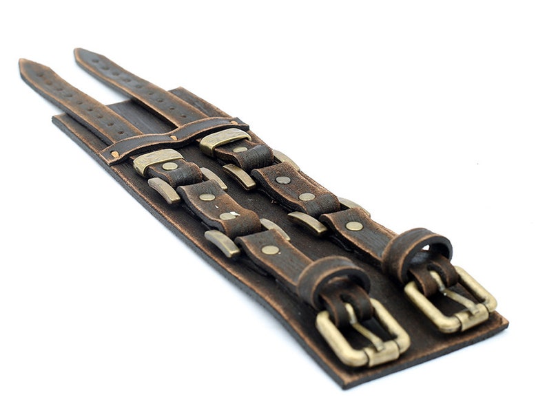 May include: A brown leather cuff bracelet with two rows of buckles and studs. The buckles are a brass color and the studs are silver.