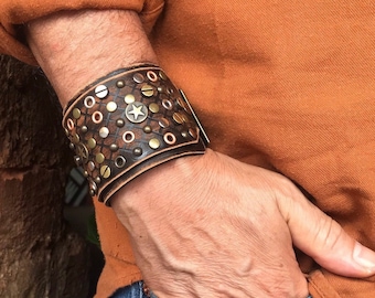 May include: A brown leather cuff bracelet with metal studs and a star design. The bracelet is worn on a person's wrist.