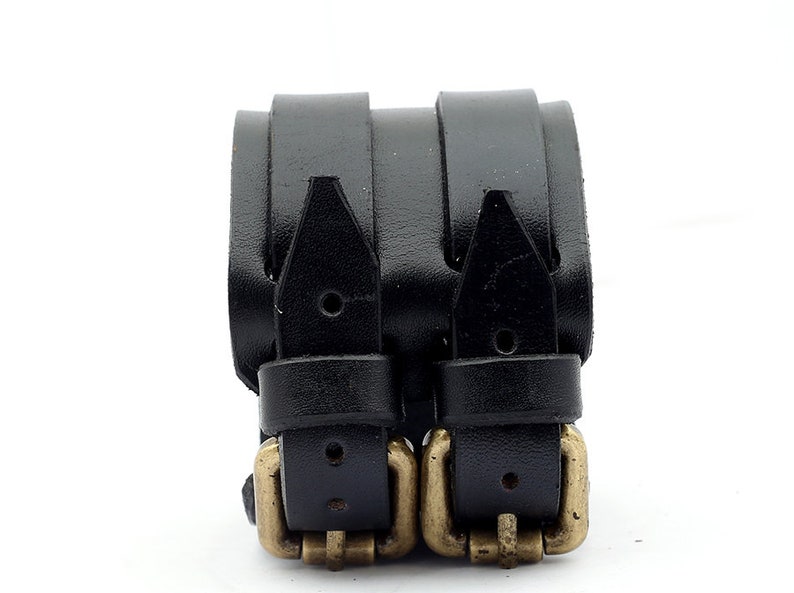 Puede incluir: Un brazalete de cuero negro con doble hebilla y detalles dorados.