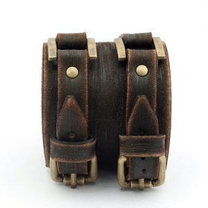 May include: A brown leather cuff bracelet with two buckles and brass accents. The bracelet has a distressed look.