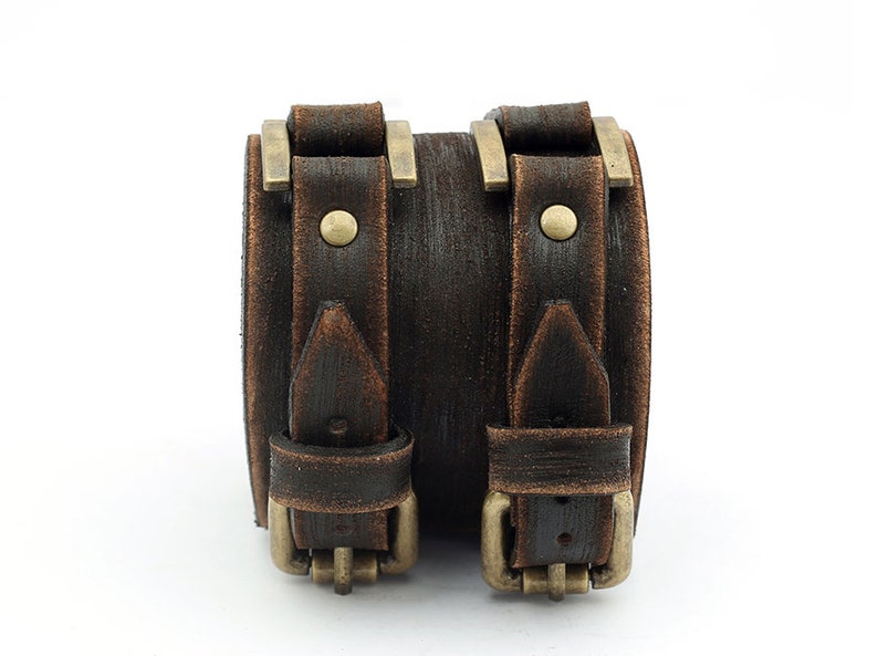 May include: A brown leather cuff bracelet with two buckles and brass accents. The bracelet has a distressed look.