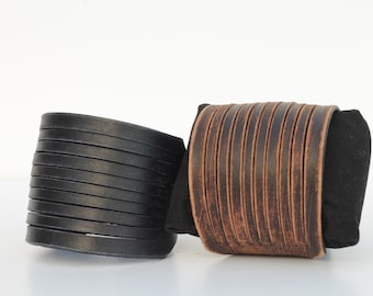 May include: Two leather wrist cuffs. One is black, the other is brown. Both cuffs are wide and feature multiple horizontal strips of leather. The brown cuff has a distressed appearance. The cuffs are displayed on a white surface.
