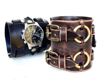 May include: Two leather wrist cuffs. One is black with a silver watch face. The other is brown with brass-colored buckles and rings. The watch has a black and silver face with multiple dials and a black leather band.