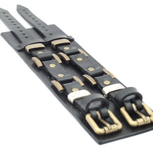 May include: Two black leather bracelets with brass buckles and studs. The bracelets are layered on top of each other.