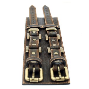 May include: A brown leather cuff bracelet with two buckles and metal accents.
