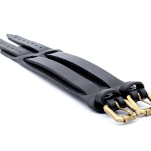 Puede incluir: Un brazalete de cuero negro con cierre de triple hebilla. Las hebillas son de lat&oacute;n y tienen un acabado pulido.