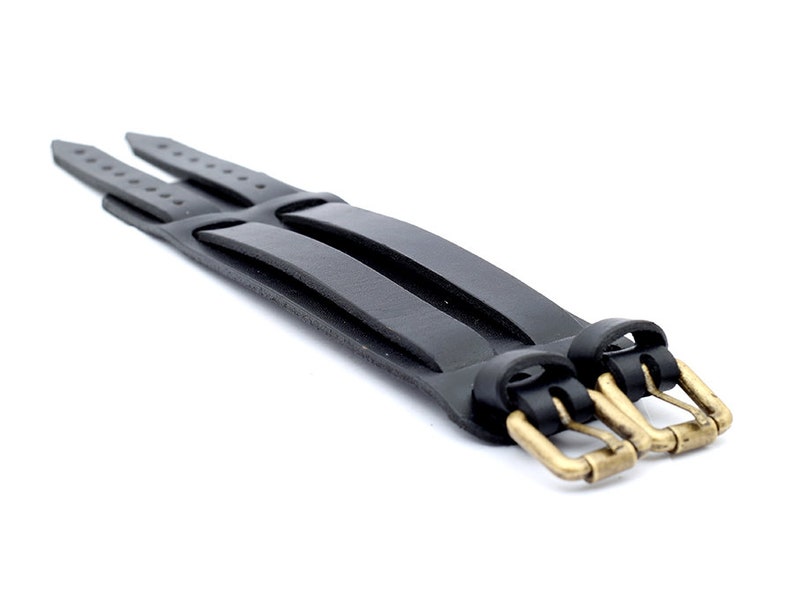 Puede incluir: Un brazalete de cuero negro con cierre de triple hebilla. Las hebillas son de lat&oacute;n y tienen un acabado pulido.
