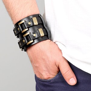 May include: A black leather cuff bracelet with gold-toned metal accents and rivets. The bracelet is wide and has a layered design.