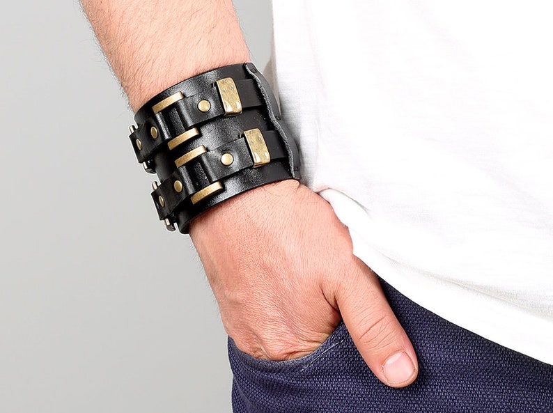May include: A black leather cuff bracelet with gold-toned metal accents and rivets. The bracelet is wide and has a layered design.