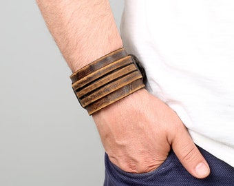 May include: A brown leather cuff bracelet with three horizontal straps. The bracelet is worn on a person's wrist.