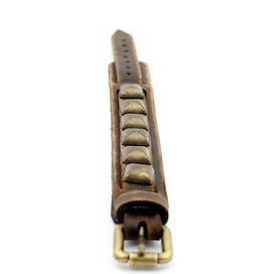 Può includere: Un braccialetto in pelle marrone con una fibbia in ottone e borchie a forma di piramide.