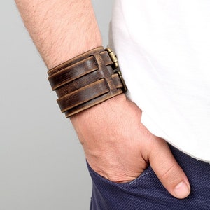 Puede incluir: Un brazalete de cuero marr&oacute;n con una hebilla de lat&oacute;n. El brazalete se lleva en la mu&ntilde;eca de una persona.