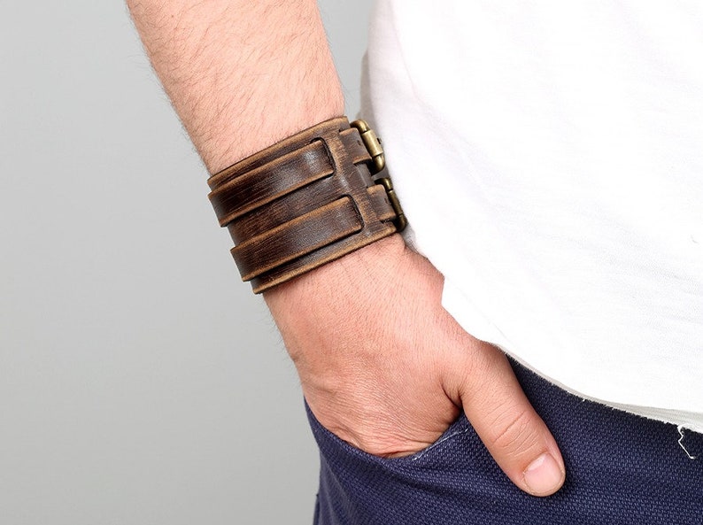Puede incluir: Un brazalete de cuero marr&oacute;n con una hebilla de lat&oacute;n. El brazalete se lleva en la mu&ntilde;eca de una persona.