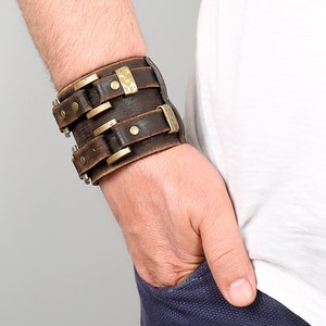 May include: A brown leather cuff bracelet with brass accents and rivets. The bracelet is adjustable and has a wide, flat design.