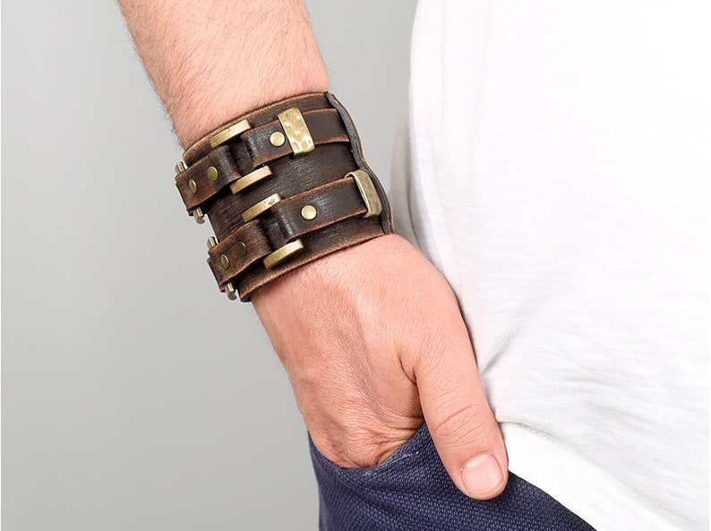 May include: A brown leather cuff bracelet with brass accents and rivets. The bracelet is adjustable and has a wide, flat design.