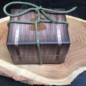 May include: A wooden box with a brown and tan woodgrain pattern, tied with green rope. The box is sitting on a wooden slice.