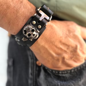 May include: A black leather bracelet with a silver skull charm and silver studs. The bracelet has a buckle closure.