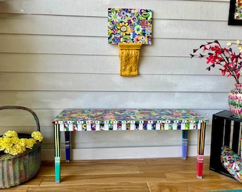 May include: A colorful bench with a floral pattern top and multi-colored legs. The bench is surrounded by decorative items, including a basket of yellow flowers, a vase with red flowers, and a wall-mounted floral artwork.