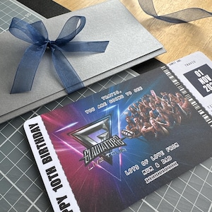 K&ouml;nnte beinhalten: Eine silberne Geschenkkarte mit einer marineblauen Schleife und ein Konzertticket f&uuml;r die "Gladiators Live Tour" mit dem Text "Happy 10th Birthday". Auf dem Ticket steht auch "Lots of love from Mum & Dad".