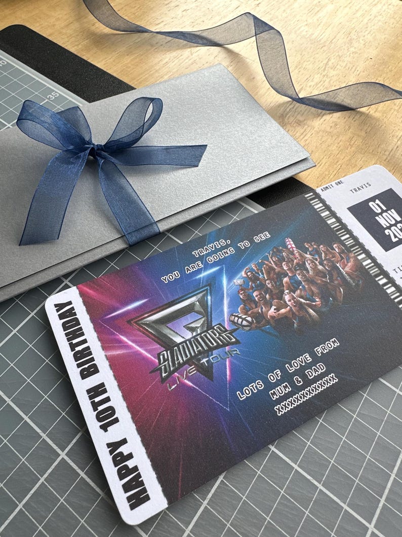 K&ouml;nnte beinhalten: Eine silberne Geschenkkarte mit einer marineblauen Schleife und ein Konzertticket f&uuml;r die "Gladiators Live Tour" mit dem Text "Happy 10th Birthday". Auf dem Ticket steht auch "Lots of love from Mum & Dad".