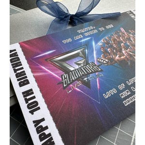 K&ouml;nnte beinhalten: Eine graue Geschenkbox mit einer marineblauen Schleife. Die Geschenkkarte tr&auml;gt die Aufschrift "HAPPY 10TH BIRTHDAY" und "TRAVIS, YOU ARE GOING TO SEE" mit dem Logo "GLADIATORS LIVE TOUR". Die Karte hat einen rosa-blauen Farbverlauf.