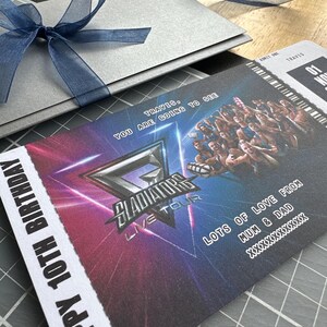 K&ouml;nnte beinhalten: Ein silberner Geschenkumschlag, der mit einem blauen Band gebunden ist, neben einem Konzertticket. Auf dem Ticket steht "Gladiators Live Tour" und "Happy 10th Birthday". Das Ticket hat ein farbenfrohes Design mit dem Text "Travis, You are going to see".