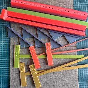 May include: A collection of multi-colored plastic crafting tools including a long red perforated bar, a green bar, four grey triangular corner guides, and several T-shaped gauges labeled 2mm, 4mm, 6mm, 8mm, and 10mm, along with thin strips.