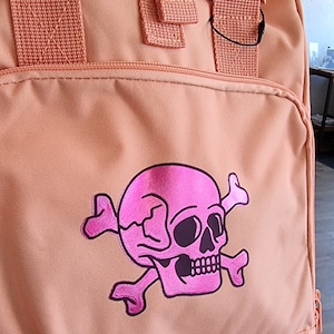 Może przedstawiać: Różowy plecak w kolorze brzoskwiniowym z nadrukiem różowej czaszki i skrzyżowanych kości. Czaszka jest obwiedziona czernią i ma różowy blask.