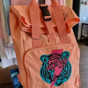 Op de afbeelding: Een perzikkleurige rugzak met een tijgerprint en een zwarte bliksem. De rugzak heeft een zwarte gesp en een zwarte band. De tekst "Je Paris" is gedrukt op de zijkant van de rugzak.