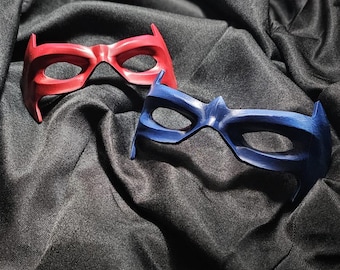 Peut inclure: Deux masques de style super-héros, un rouge et un bleu. Les deux masques ont un design similaire avec des ouvertures pour les yeux et des bords pointus. Ils sont présentés sur un tissu sombre et texturé.