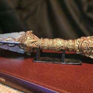 Puede incluir: Una daga metálica con hoja triangular y un mango ornamentado que presenta un rostro esculpido en el pomo. La daga descansa sobre un pequeño soporte negro encima de un libro rojo oscuro con el texto "Histoire universelle" en el lomo.