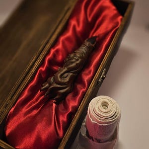 Puede incluir: Una varita tallada de color marrón oscuro descansa dentro de una caja de madera forrada con satén rojo. La varita es el punto focal, con tallas detalladas. Una venda enrollada está en primer plano.