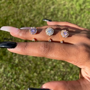 May include: A set of four gold rings with clear and purple gemstones. The rings are worn on a finger with black nail polish.