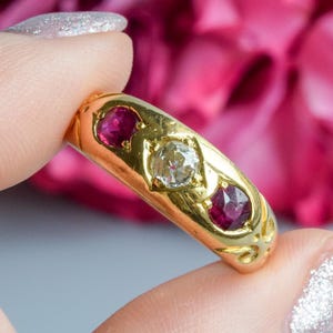 Puede incluir: Un anillo de oro con un diamante central y dos rubíes rojos. El anillo se presenta sobre un fondo de flores rosas y moradas. El diseño del anillo es ornamentado.