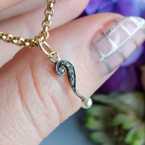 May include: A gold chain necklace with a decorative pendant. The pendant is black with a swirl design and small, clear stones, and a tiny pearl. The necklace is held against a backdrop of purple and peach flowers.