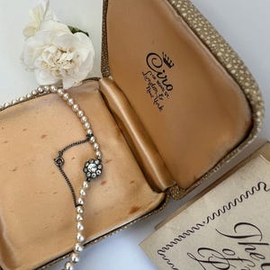 May include: A string of cream-colored pearls with a central flower-shaped clasp featuring clear rhinestones, housed in a textured tan jewelry box. The interior lid is printed with the text Ciro of Bond St. London & New York with a crown icon.